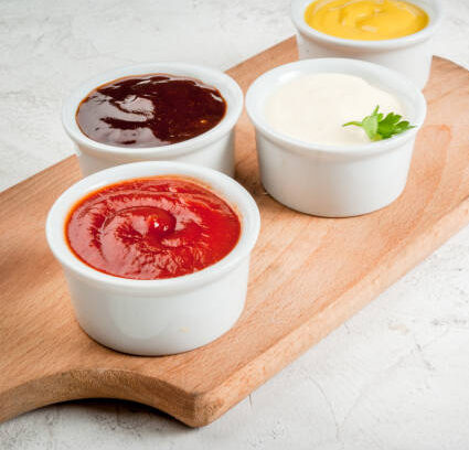 Classic set of sauces in white saucers: American yellow mustard, ketchup, barbecue sauce, mayonnaise. On cutting board white stone concrete table close view, copy space